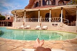 Image shows a pair of feet, resting on a lounger in front of a kidney-shaped pool, four empty loungers and a deck across the way.