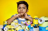 Wide-eyed young boy against a yellow background with a pile of minion toys in front of him.