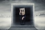 A man sat in a concrete box with a paper bag over his head displaying a drawn sad face.