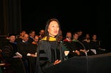 Woman dressed in academic regalia speaking at lectern with two seated rows of similarly dressed professors behind her.