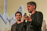 A closeup photo of moderator Carlos Tejera to the left of  director Marcel Barrena as he answers audience questions about his latest feature film, Mediterraneo: The Law of the Sea. Marcel holds a microphone in his right hand as he gestures to the audience with his left. Behind them is a projection of Little Havana’s Tower Theater marquee.