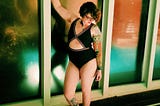 A woman with short curly brown hair posing in a doorway wearing a swimsuit.