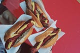 Three hot dogs on buns, held with white paper over a red background