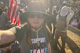 The author is standing in front of a large crowd of people protesting. He’s wearing a hat and sunglasses. He has long brown hair and a beard. His t-shirt says “I support Black trans futures.” There’s an American flag behind him on the left and several protest signs. One sign says, “stop lying”. A person in the background is wearing a trans flag on the right. Another person in the background is waving a Mexican flag.
