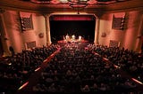 Photograph of the interior space of the historic Lobero Theatre, overlooking the audience from the back of the venue towards the stage. There are approximately 600 patrons in the venue, taking up every seat in the audience. A band is playing on the stage, featuring electric guitars, an upright bass, and a cajón. The lighting is dim with subtle, warm colors, conveying a classy atmosphere.