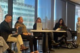 Estefania Cubillos Nova sitting on a panel with four other people. She is wearing black pants, a brown blazer and her hair is tied back. In front of her is a desk with a laptop.