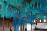 An industrial warehouse space, with a concrete floor and brick walls. People wander around, wearing black headsets. Above them is a rendering of a virtual entity: rows and rows of narrow slats of bright teal form a layered, undulating shape.