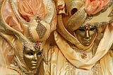 A man and woman in golden full-face masks and rich golden-and-peach-colored costumes.
