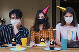 three people sit at a table with party hats and masks on. the table has cupcakes, NYU Local logos cover the candles.