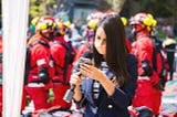 Female news anchor checks her phone, holds microphone, firefighters in background: by Mike Ramirez Mx on Pixabay
