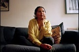 [Image ID: Feminist leader bell hooks on a couch with her chin resting on her hand, wearing a yellow shirt and looking thoughtful, per usual.]