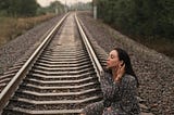 Alt-text: A woman in dark clothing sits on a railway track at dusk, symbolizing crossings between danger and freedom.