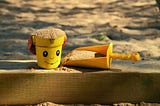 A sand box with a yellow bucket, that shows a smiling face, and a shovel next to it.