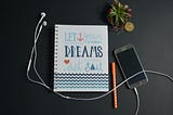 A mobile phone with attached headphones accompanied by notebook with title that reads “Let Your Dreams Set Sail.”