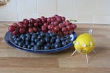 Lemon pig — standing on cocktail stick legs, with eyes of cake candle holders and a curly tail of tinfoil, bearing a coin in its mouth, beside a plate of red grapes and blueberries.