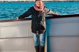 A woman stands on the deck of a boat. Water stretches behind her. Her arms are resting on the siderails.