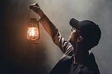 A man in a black ballcap holding a lit lantern up towards the darkness.