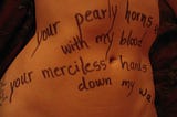 A woman’s belly with words written in ink pen on her skin, “your pearly horns, with my blood, your merciless hands, down my walls.”