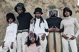 Six Black people standing outside of a rock formation wearing 80s style fashion