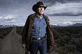 The television character Walt Longmire in blue denim shirt and jeans wearing a cowboy hat and leather coat holding a rifle. Mountains and a gravel road in the background with a dramatic, storm laden sky above.