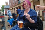 Claire sat in her electric wheelchair, with someone else's pint of beer in her chair's cupholder. Someone is handing her a gin and tonic.