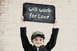 Photo of a boy holding up a small chalkboard. On the board is written the words, “Will work for love.”