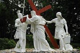 An outdoor Station of the Cross depicts Jesus carved out of stone, struggling to carry a wooden cross surrounded by statues of Roman centurions.
