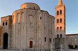 A large, circular church structure of beige stone, with a tall bell tower nearby; upper levels catch the golden light of sunset.