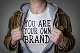 Man holding jacket open to show a t-shirt that says “You are your own brand”