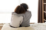 Mother and daughter sit on the bed, the girl’s head leaned against her mom