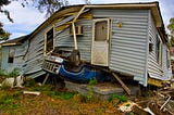 An unusual scene, with a damaged and twisted house resting on top of a blue vehicle that is upside down.