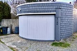 A round fronted, stand-alone garage with a curved rolling door and shingled sides facing an alley.