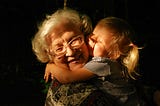 Grandma hugging blonde haired two year old girl on dark background with light shining on their faces.