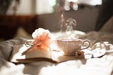 ceramic teacup with steaming coming out of it on open notebook beside pink flower pen on bedside