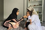 In the image, two women and a young child are seated on a patterned carpet indoors. One woman, dressed in a black and pink outfit with a black hijab and glasses, is gently touching the face of the child, possibly in a comforting or affectionate gesture. The child, who appears to be a toddler, is sitting on the lap of the second woman, who is wearing a white outfit and a light-colored patterned hijab. Behind them is a white shelf containing various decorative plates and items.