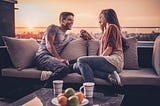 A man and a woman sit together on a roof terrance while the sun is setting. They are holding hands and smiling at each other, but it feels romantic rather than sexual.