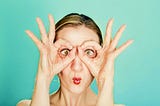 A comical portrait of a young woman with buldging eyes and pursed lips. The woman’s hands make OK signs. She raised them to her face and looks through them, as though looking through the eye-holes of a mask or binoculars.