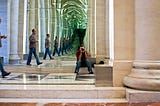 An “infinite mirror” image of a man walking into a columned hallway. Another man, seated and holding a camera to his eye, faces the viewer,