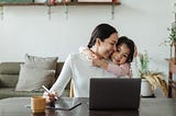A woman working from home while hugging her daughter.