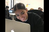 Fazia Bouheaoura, a web developer smiling graciously, sitting in of front of her laptop working in a room filled with women.