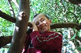 The author as a child clinging onto a tree branch. He’s wearing a red sweater, has brown hair, and is smiling at the camera. In the background, lots of tree branches and green leaves are visible.