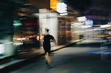 Nighttime city street with bright storefront lights and colorful neon signs blurred by motion. A single person in dark athletic clothing and a cap runs away from the camera along the sidewalk, captured with intentional motion blur. Reflections and streaks of light create a dynamic, fast-paced scene with soft, abstract edges and a sense of speed.