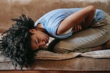 A female with skin the color of coffee with a single-cream, wearing a baby blue acrylic vest and beige pants is kneeling on a brown velour sofa. She’s hunched over in pain, clutching her stomach. Her medium length, dark brown hair is wavy and messy. Her left ear is exposed. Her right temple is touching the sofa. The remaining right side of her face is slightly lifted from the sofa.