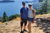 A man and woman standing on a rocky viewpoint in summer with a backdrop of ocean and trees.