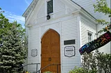 White clapboard church with oak doors, stained glass windows, and a colorful pride flag out front.