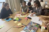 Workshop Attendees, three smiling women with brown hair sitting at a table with paper and colorful post it notes. A  fourth woman whose hands are visible at the right of the image sits next to a closed laptop computer covered with stickers, a paper cup and a mobile phone. Pens and yellow sticky notepads are scattered on the table top.