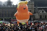 Giant Trump diaper baby balloon “welcoming” Trump to London, balloon is 6 meters (20 feet) tall, floating just over a small crowd in front of some random London buildings, balloon depicts Trump as an angry orange baby dressed only in a diaper and holding a cell phone
