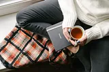 Woman wearing white sweater and jeans sitting on windowsill with notebook and cup of tea, on top of cozy blanket.