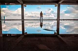 A man stands on a horizon, a bright blue sky before him and a water reflection of him and the sky underneath him.