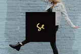 blonde woman walking making big steps ahead of a light blue brick wall, in the foreground is the album cover of Sylvan Esso, by duo Sylvan Esso